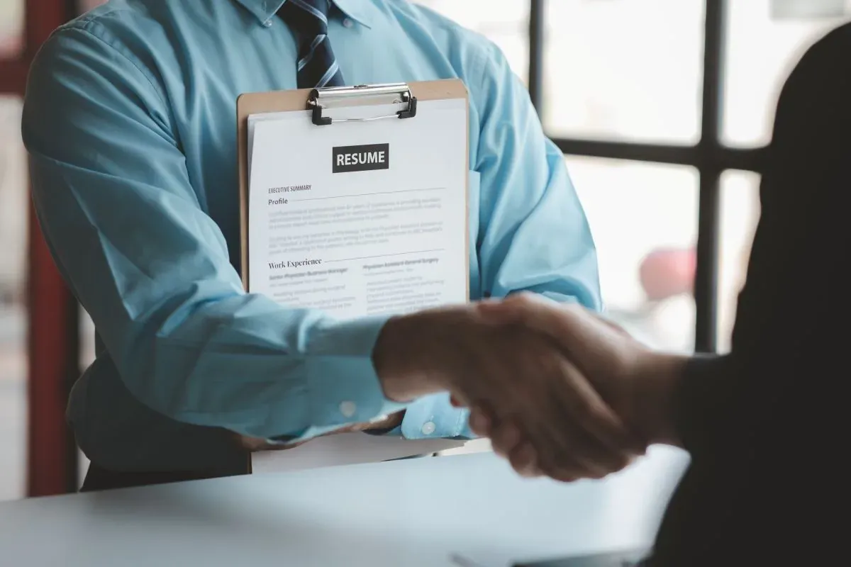 Candidate holding a resume on a clipboard shakes hands with an interviewer across a desk